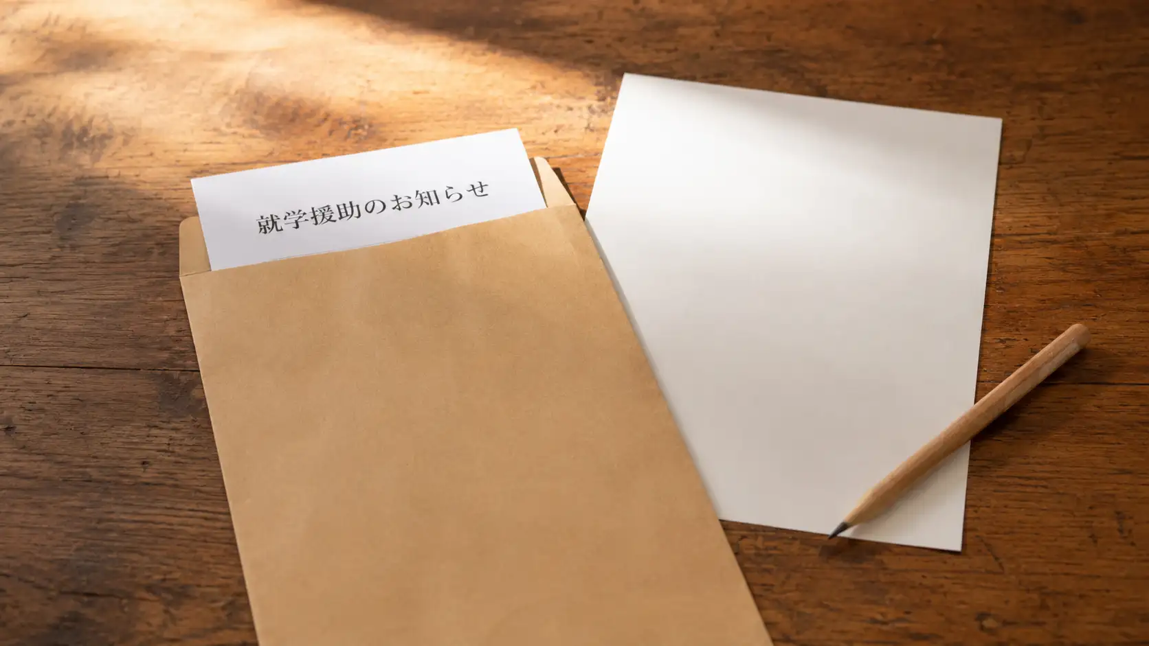 木の机に置かれた茶色い封筒と就学援助の申請書らしき用紙、鉛筆